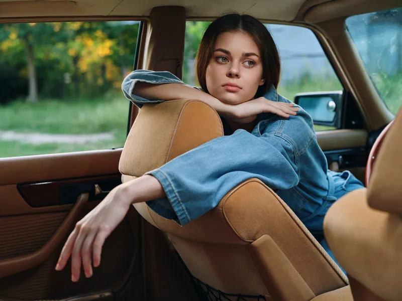 Young woman in denim jacket resting in a vintage car, capturing a moment of contemplation on a sunny day in a natural setting with greenery.