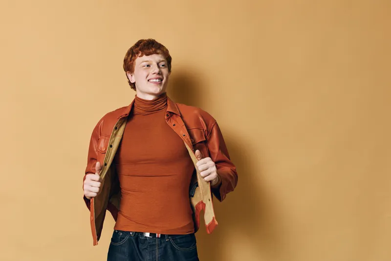 Young man with red hair wearing orange jacket and brown turtleneck posing smiling against yellow background. Casual style, confident expression and relaxed posture in studio interior.