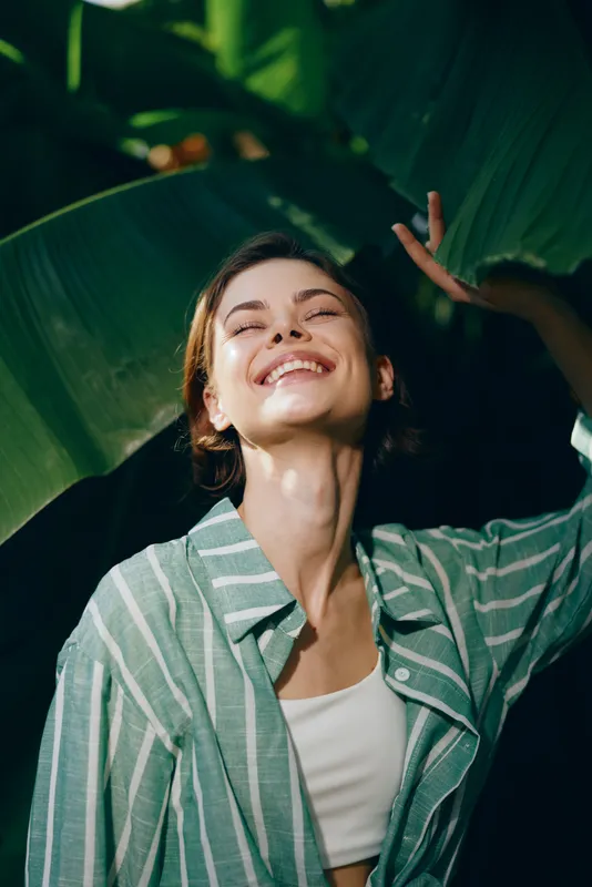 Joyful woman basking in warm sunlight beneath lush tropical leaves, wearing a striped shirt, radiating carefree summer energy.