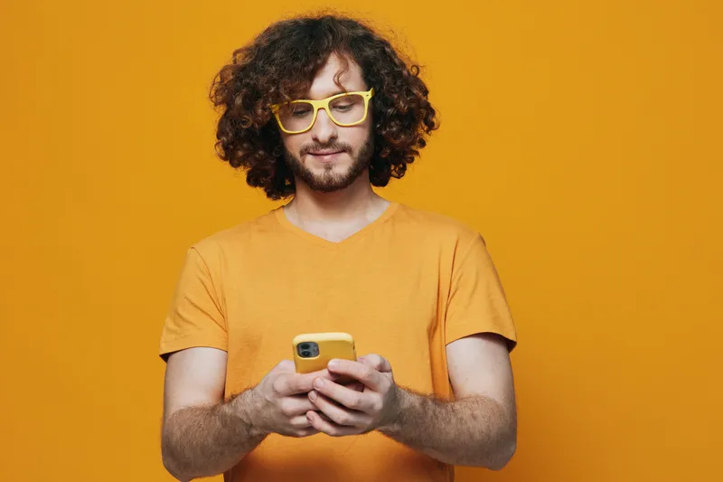 Stylish young man with curly hair and glasses using smartphone against a vibrant yellow background, embodying modern lifestyle and digital connection
