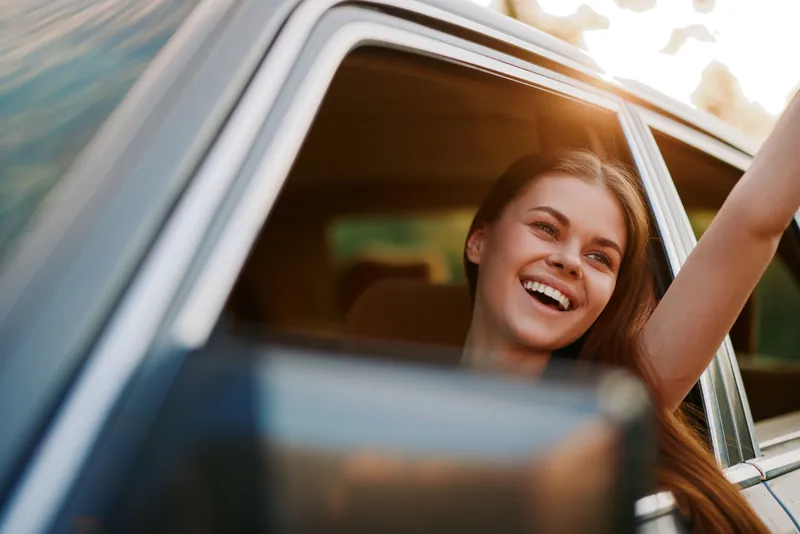 Happy young woman enjoying a road trip with arms raised from a car window during sunset, embodying freedom and joy in nature.