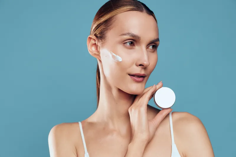 Young woman with clean skin applies white cream on her face holds cosmetic jar looks away stands against blue background skincare beauty concept