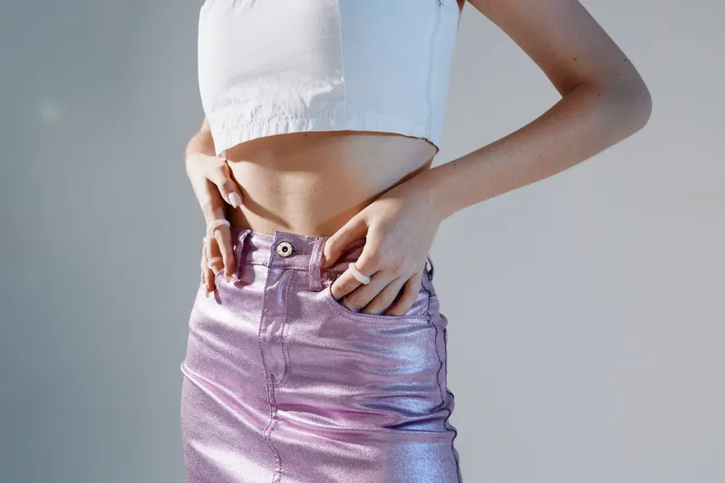 Woman beauty fashion concept with a close-up of a female model wearing a shiny purple skirt and a white crop top. Studio lighting highlights the texture and style.