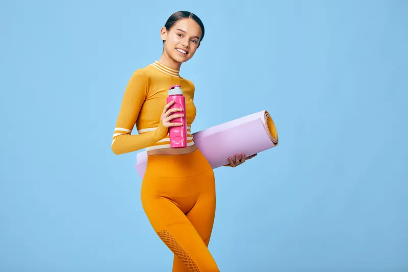 Woman fitness model in bright orange athletic wear, holding a water bottle and a yoga mat, standing against a blue backdrop, ready for workout, wellness and active lifestyle.