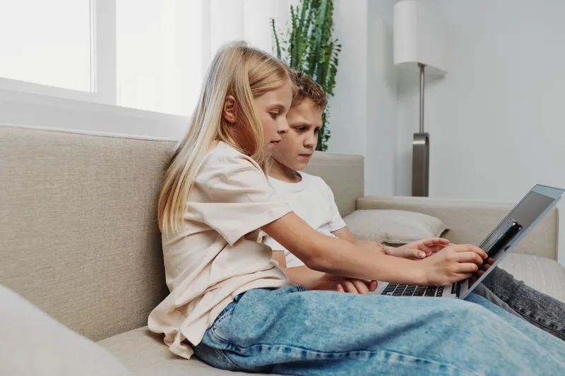 Two focused children sitting on a beige sofa using a laptop together in a bright living room. Concept of learning, technology, cooperation, education, and modern childhood activities.