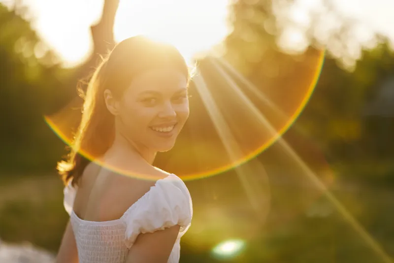 Young woman smiling in sunlight, wearing a white dress, surrounded by nature, conveying joy and serenity during golden hour