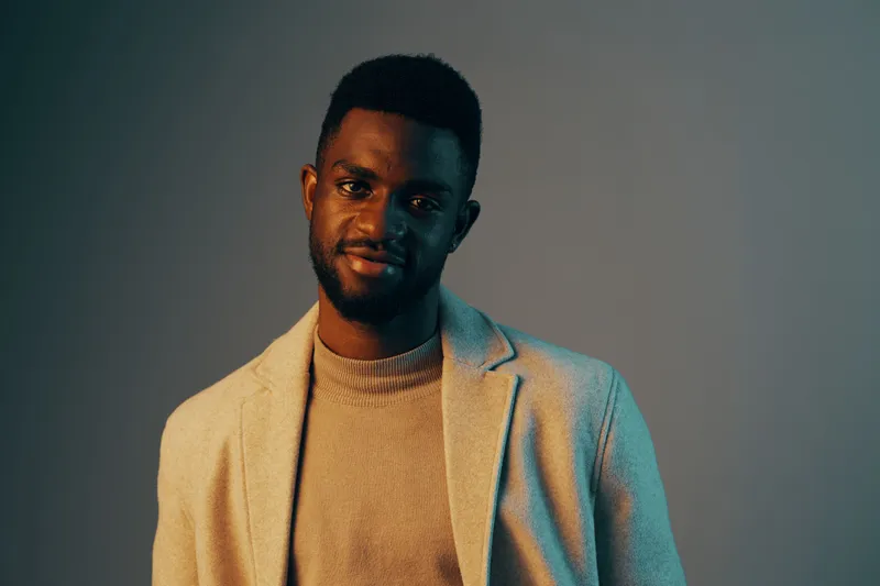 A stylish young African man wearing a beige blazer and a light sweater smiles confidently against a soft blue backdrop, exuding modern elegance and charm