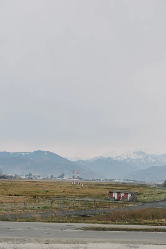 Rural landscape with dry grass and small red house under cloudy sky. Mountains visible in background create calm and isolated atmosphere.