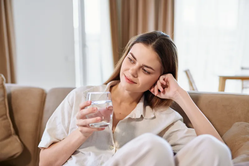 Woman holding glass of water on sofa at home, relaxed portrait of a casual young female drinking and smiling, hydration and wellness in cozy living room with natural light.