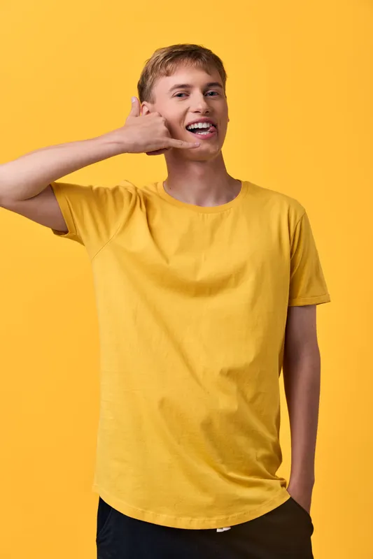 Cheerful young man in a yellow shirt makes a phone hand gesture against a bright yellow backdrop, conveying casual communication, friendly energy, and modern lifestyle in a studio setting.