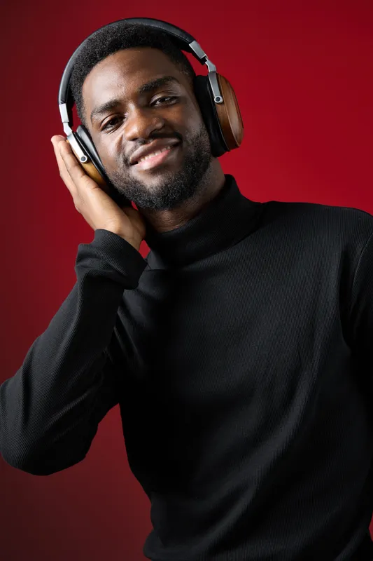 Young African man in a black turtleneck enjoying music with headphones against a red background, conveying happiness and relaxation through his smile