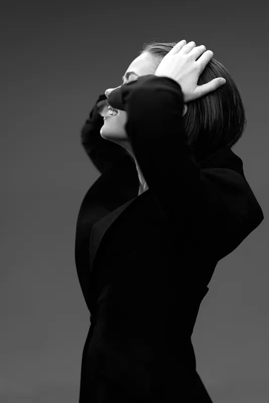 Black and white portrait of a thoughtful woman in a dark jacket posing with closed eyes and hands in her hair.
