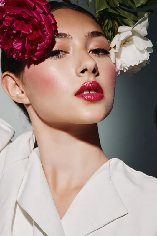 Close-up of a young woman with glossy red lips and flawless skin wearing bold flower accessories and a stylish white outfit. Concept of beauty, fashion, and creative editorial makeup.
