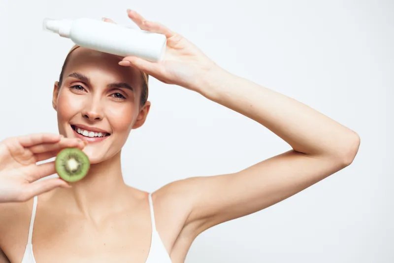 Cheerful woman applying sunscreen, holding kiwi slice, bright studio light, fresh skincare moment, sun protection vibe, healthy glow, casual pose, white background, natural colors