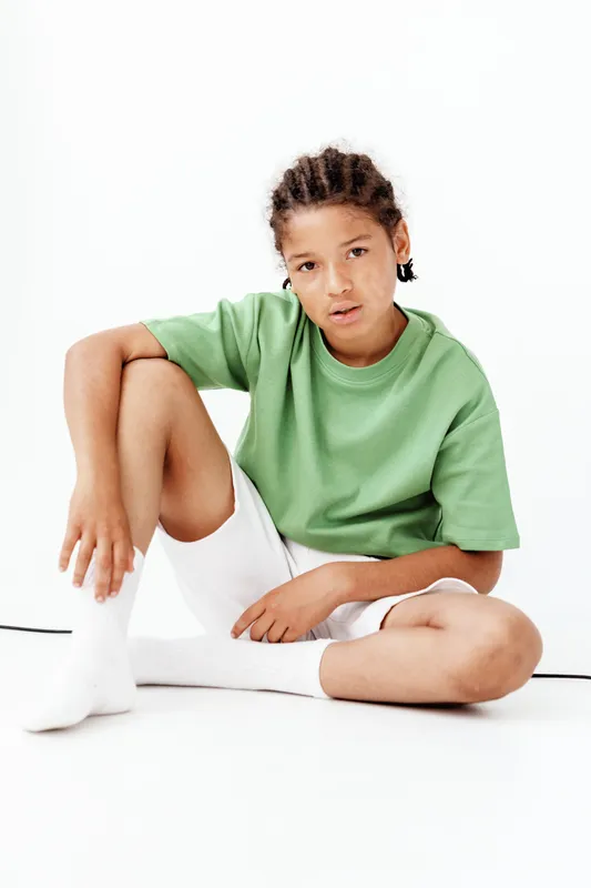 Мальчик в стильной одежде сидит на полу в белых носках и зеленой футболке, излучая крутую и непринужденную атмосферу