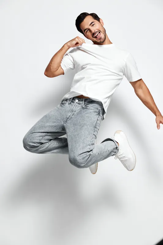 Happy smiling young man in casual white t-shirt jumps with excitement while posing for the camera against a plain light background. Joyful lifestyle concept.
