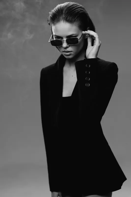 Stylish woman wearing sunglasses and black blazer in a smoky studio setting, black and white portrait with a confident expression