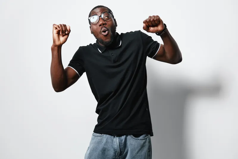 African American man with dark skin wearing glasses and black polo shirt expressing excitement with raised fists on white background. Casual style and joyful emotion.