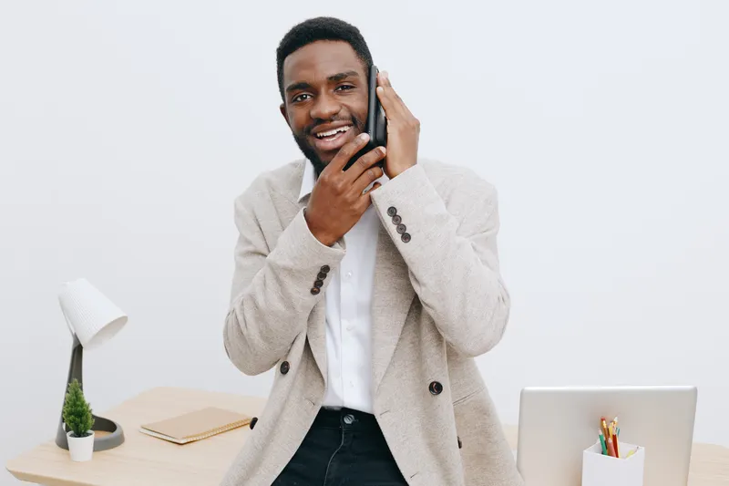 Professional young African American man in stylish beige blazer smiling while talking on a mobile phone in modern office interior with workspace accessories, background, and neutral colors. People lif