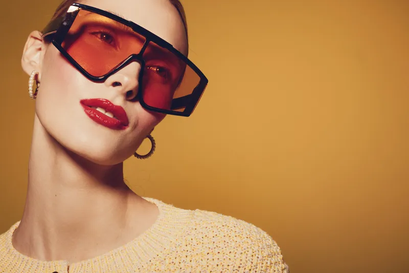 Stylish woman with oversized red sunglasses and bold red lipstick poses against a warm yellow background. Fashion concept with modern accessories and confident attitude.