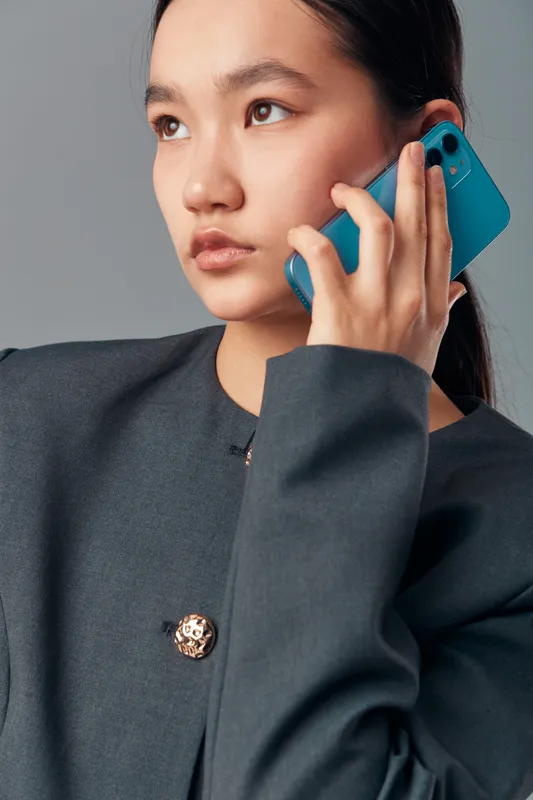Young woman holding blue smartphone near ear in dark jacket for communication with thoughtful expression on gray background. Technology, conversation and portrait closeup.