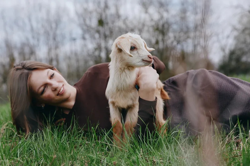 A woman lies on grass beside a curious goat kid in a sunlit field, sharing a quiet lifestyle moment. The scene emphasizes gentle companionship, rural calm, and a playful bond between human and animal.