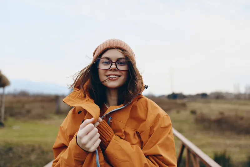 Woman lifestyle outdoors smiling portrait, orange jacket and beanie, relaxed stance by fence on rural walkway, soft daylight, green fields and distant hills