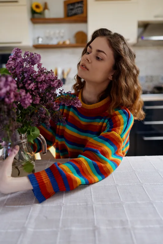 Young woman with curly hair arranges a vibrant bouquet on a white table in a sunlit kitchen, conveying a cozy home lifestyle, calm focus, gentle contentment and simple quiet rituals.