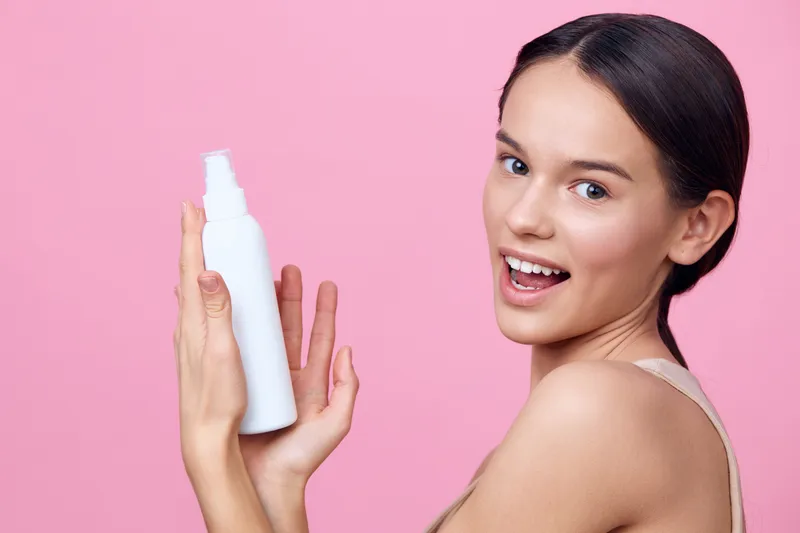 Smiling young woman holding a skincare bottle on a pink background, showcasing beauty, freshness, and self care with a playful expression