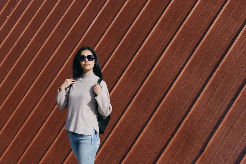 Woman in casual gray sweater and denim with backpack poses beside a diagonal wooden wall, street fashion and outdoor lifestyle in an urban setting, stylish, confident vibe