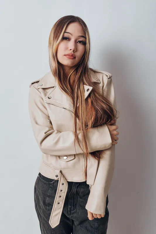 Young woman with long hair wearing beige jacket and dark pants posing with arms crossed against white background. Casual style and confident look in studio.
