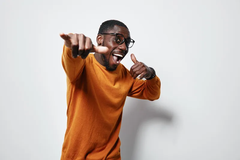 African American man with dark skin wearing glasses and orange sweater smiling and pointing at camera in studio. Happy male portrait with positive expression and casual style on white background.