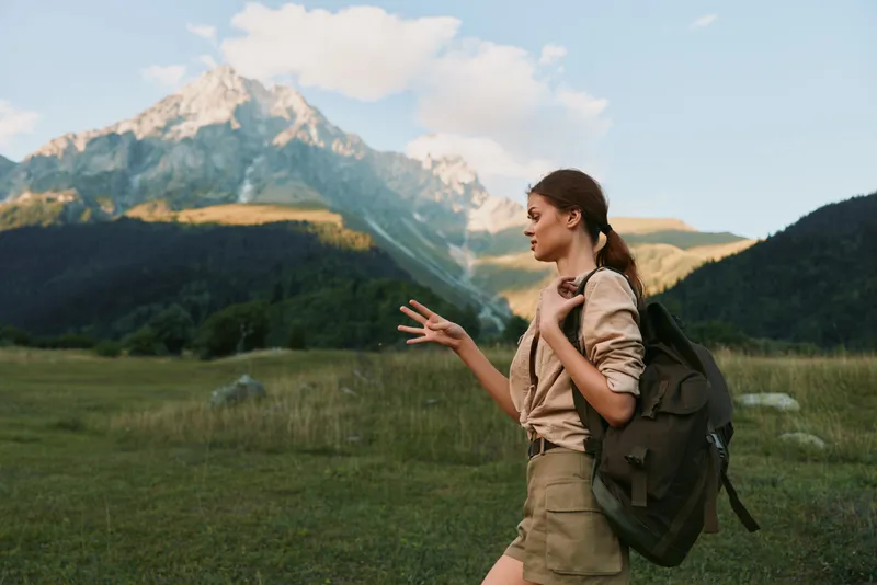 Young woman with a backpack hikes through a grassy valley beneath towering mountains, gesturing as she chats during an outdoor trek in bright daylight and fresh air.
