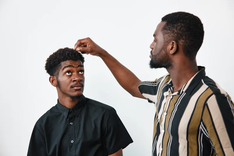Two African American men with dark skin in casual shirts interacting on white background. One man inspects the other man’s hair with focused attention and care.