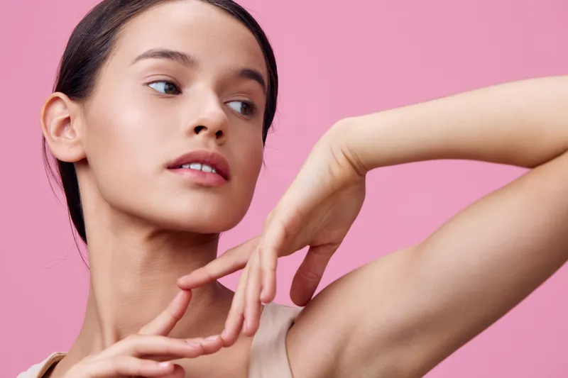 Beautiful young model with smooth skin poses gracefully against a pastel pink background, showcasing elegant hands and a serene expression for skincare concepts