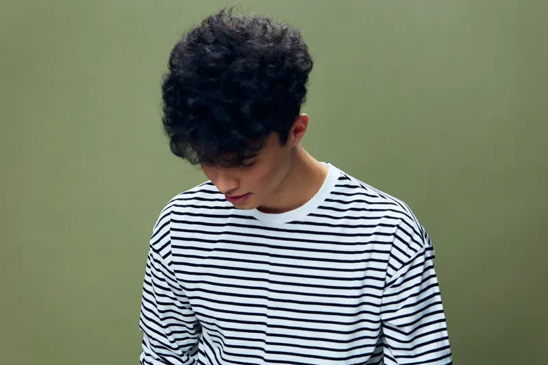 young man with curly hair wearing a striped shirt looking down against olive background, casual style, thoughtful mood, studio portrait with soft lighting