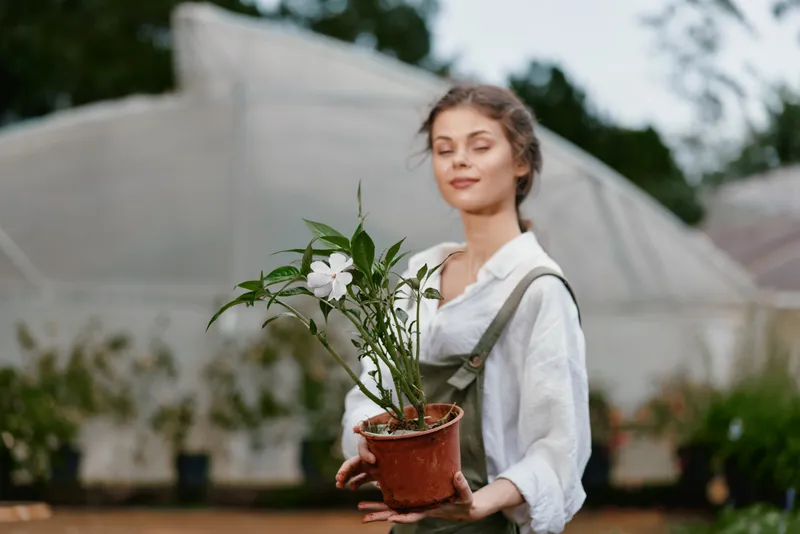 A woman with a serene expression holds a potted plant in a greenhouse, showcasing the concept of sustainable gardening and plant care