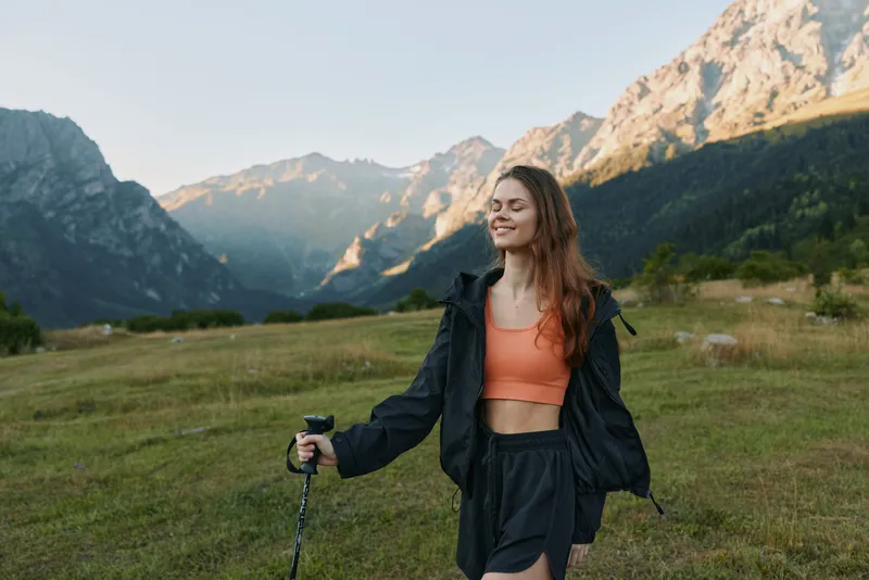 A cheerful outdoor enthusiast walks through a mountain meadow, smiling while holding a trekking pole. She wears an orange crop top and a dark jacket, radiating energy in nature.