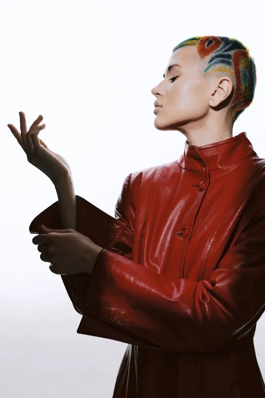 Woman with colorful shaved hair wearing a red leather jacket, posing gracefully with a profile view against a white background, showcasing edgy fashion and style.