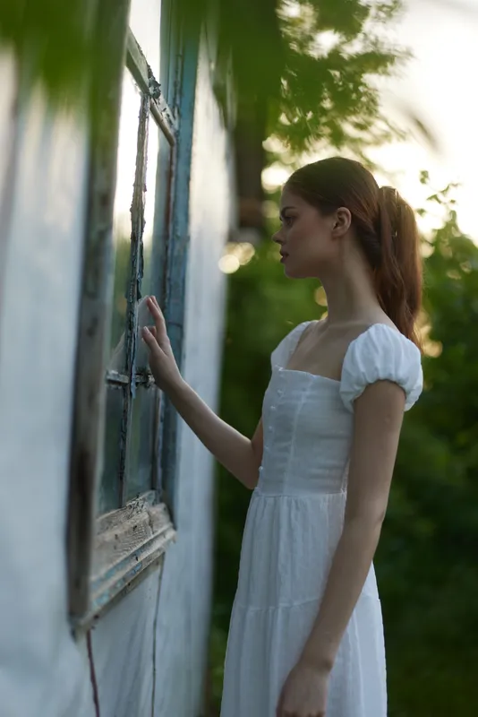 A serene young woman in a white dress stands beside a vintage window, surrounded by greenery, evoking feelings of peace and nostalgia in a sunny setting