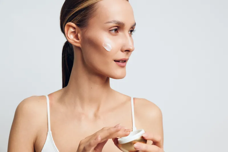 Young woman with brunette hair applies facial cream on her cheek while holding an open jar in a bright studio setting, showcasing skincare and beauty routine.