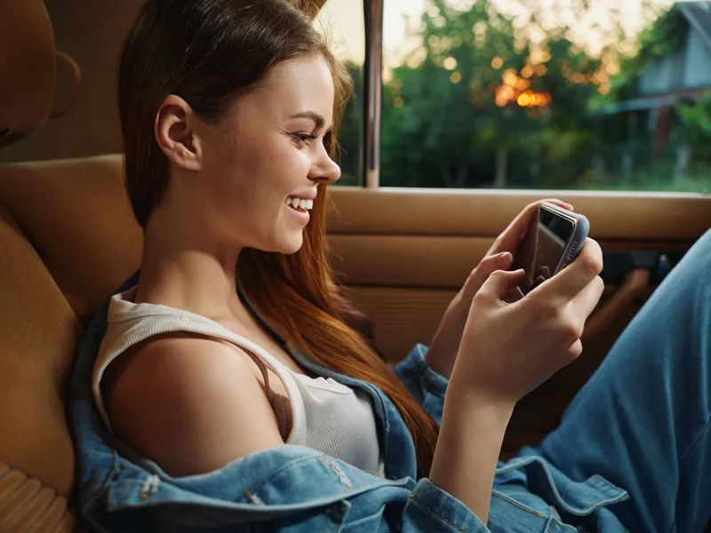 Smiling young woman using a smartphone in a car, natural lighting creates a warm atmosphere, capturing happiness, casual style, and connection.