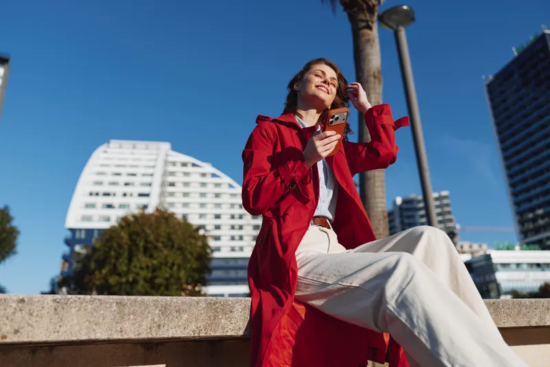 Young woman in red coat sits outdoors on stone bench holds smartphone looks up smiling enjoys sunny day urban background clear blue sky.