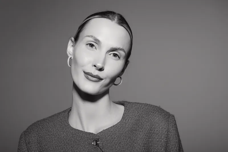 Portrait of a young Caucasian woman with pale skin and slicked back hair wearing hoop earrings and a textured top against plain background. Studio lighting.