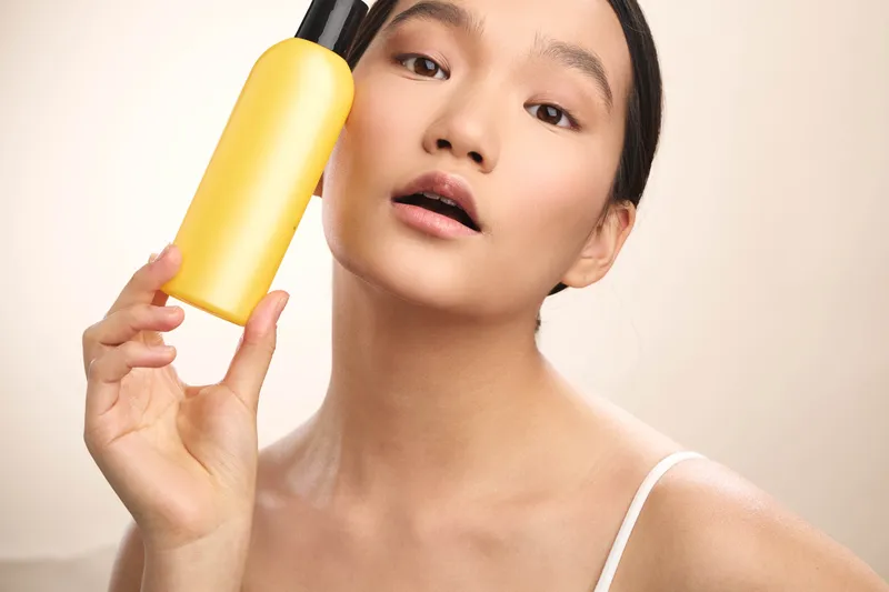 Asian woman holds a yellow bottle near her face, showcasing skincare products with a soft, confident smile in a clean, minimal setting, creating a fresh and inviting mood.