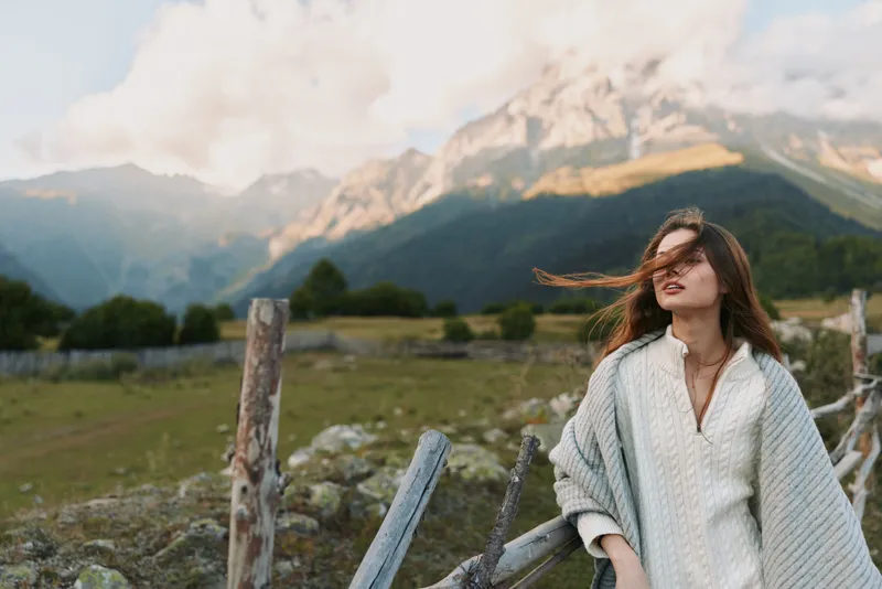 Young woman with long hair enjoys fresh mountain air while leaning on a wooden fence in a cozy knit sweater during a calm autumn day.