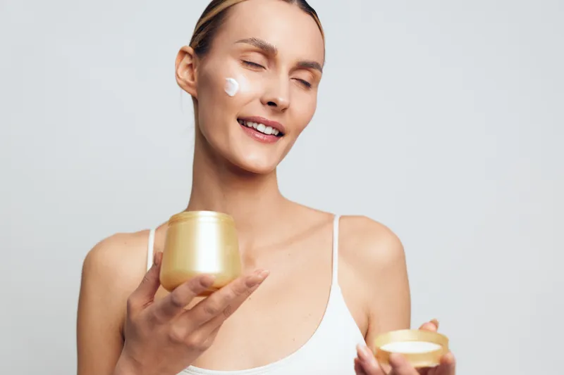 Young woman applies face cream with closed eyes holds jar of moisturizer in hand wears white tank top isolated on gray background skincare beauty concept