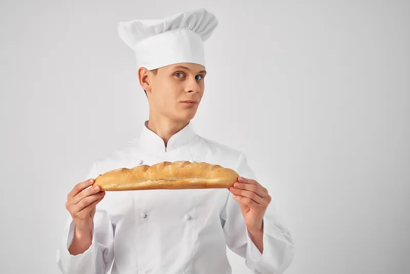 Весёлый пекарь-мужчина откусывает батон, занимаясь приготовлением еды в пекарне