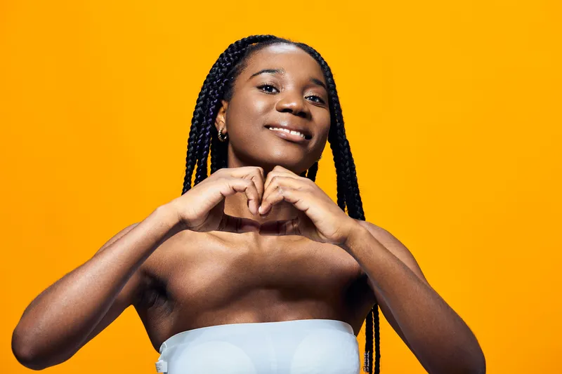 Young woman with braided hair making a heart shape gesture against a vibrant yellow background, expressing joy and love, ideal for seasonal greetings and wellness themes