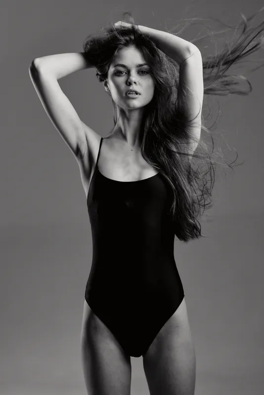Black and white portrait of a young woman in a black swimsuit with flowing hair and confident expression.